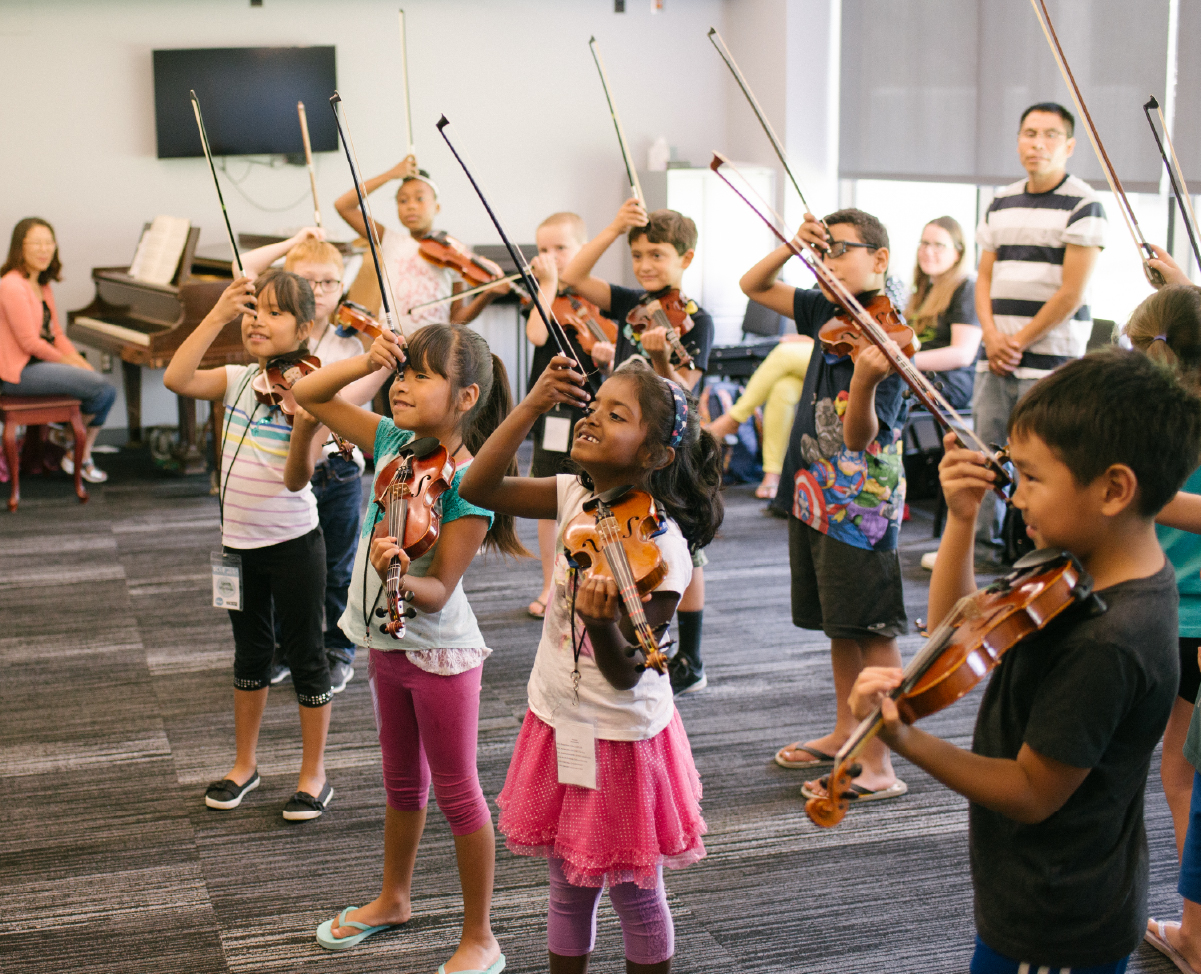 A group of young strings players humorously hold their bows to their foreheads, imitating unicorns.