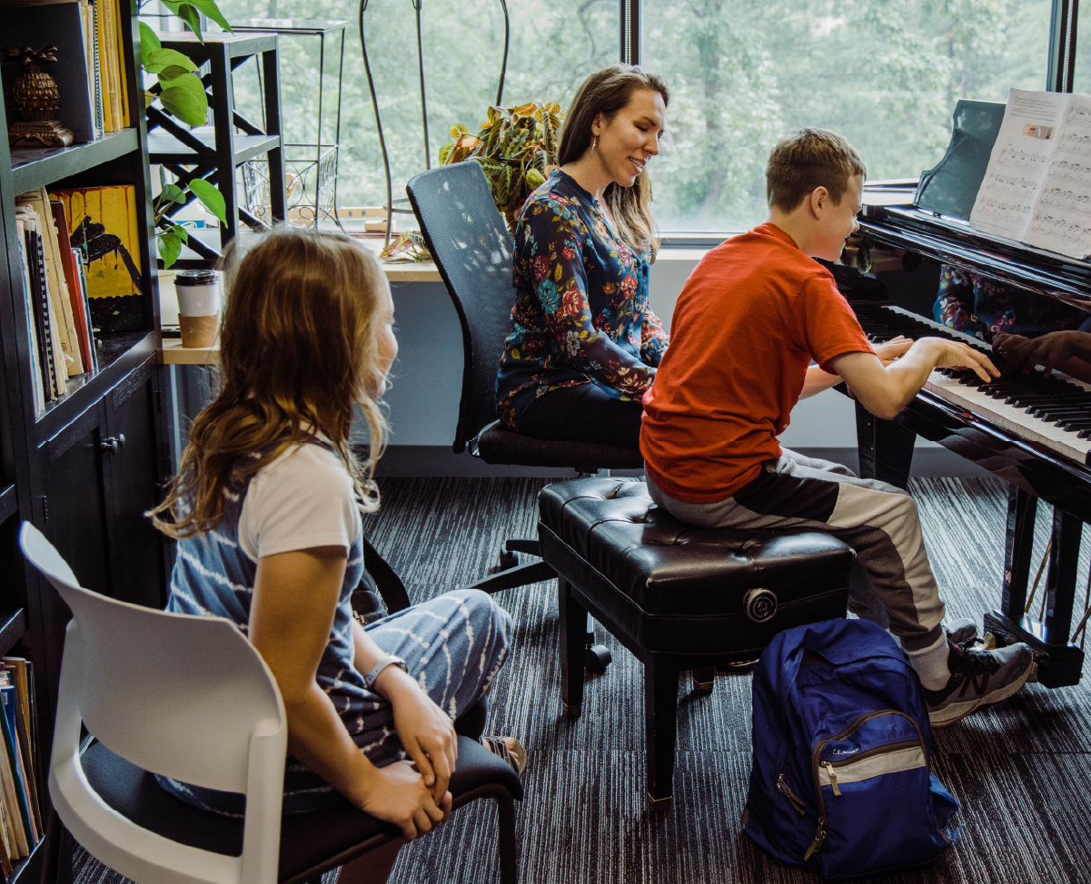 A piano teacher sits next to a young student as he plays; his sister watches from behind.