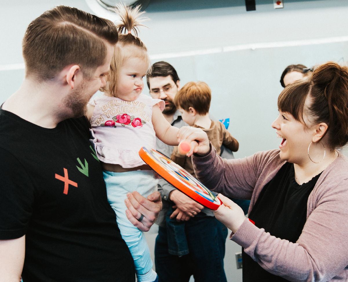 Tuneful Tots instructor Candace Jorgensen holds a drum up to a Tuneful Tots student, who is making a humorous face while being held by her father.